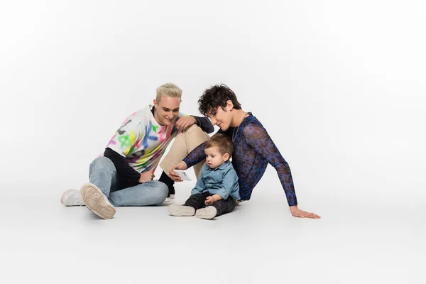 Happy gay men sitting with toddler kid and taking selfie on smartphone on grey background — Stockfoto