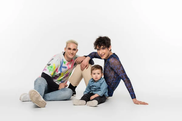 Stylish gay partners sitting near toddler son on grey background — Stockfoto