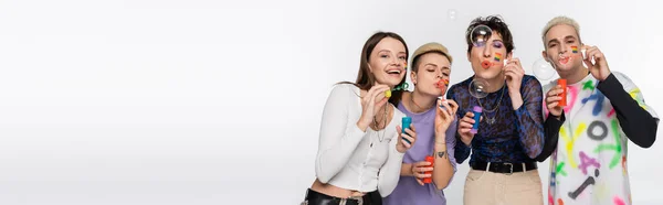 Stylish people of different identity and with lgbtq flags on faces blowing soap bubbles isolated on grey, banner - foto de stock