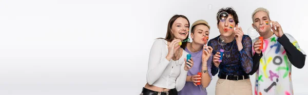 Stylish people of different identity and with lgbtq flags on faces blowing soap bubbles isolated on grey, banner — Fotografia de Stock