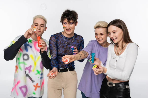 Excited lgbtq community friends in stylish clothes blowing soap bubbles isolated on grey - foto de stock