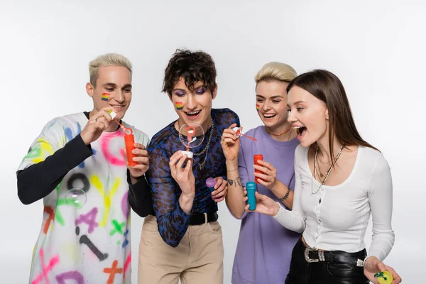 Happy diverse identity friends with lgbtq flags on faces blowing soap bubbles isolated on grey — Stockfoto