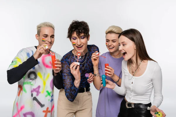 Happy diverse identity friends with lgbtq flags on faces blowing soap bubbles isolated on grey — Fotografia de Stock