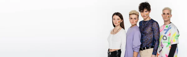 Trendy lgbtq community people with rainbow flags on faces looking at camera isolated on grey, banner — Stockfoto