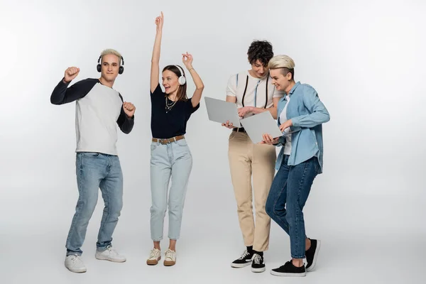 Cheerful couple dancing in headphones near friends with laptops on grey background — стоковое фото