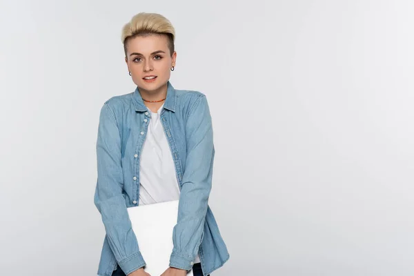 Blonde woman in denim shirt holding laptop and looking at camera isolated on grey — Stockfoto