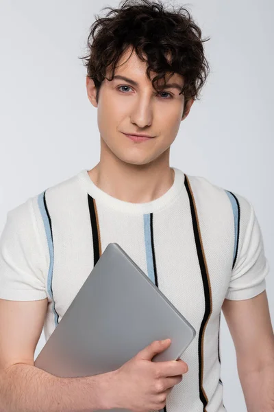 Trendy brunette man in striped t-shirt holding laptop and looking at camera isolated on grey — Foto stock