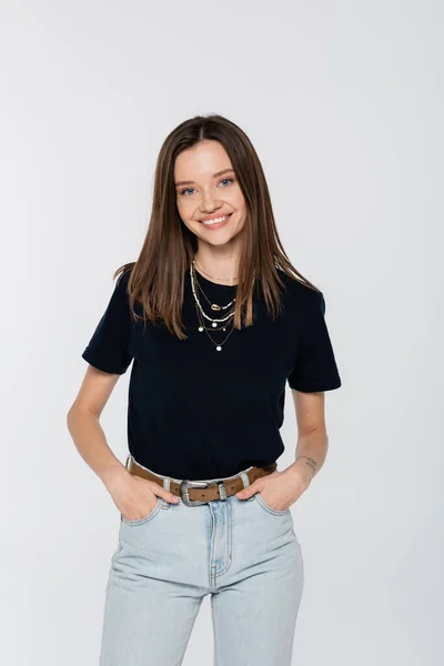 Happy woman in black t-shirt and necklaces holding hands in pockets of jeans isolated on grey — Stockfoto