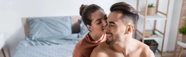 Молода жінка цілує без сорочки усміхненого чоловіка в розмитій спальні, банер — стокове фото