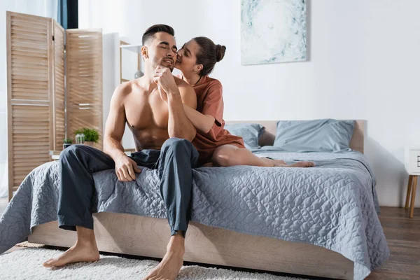 Sexy woman in t-shirt kissing shirtless man sitting on bed in pajama pants — Stock Photo