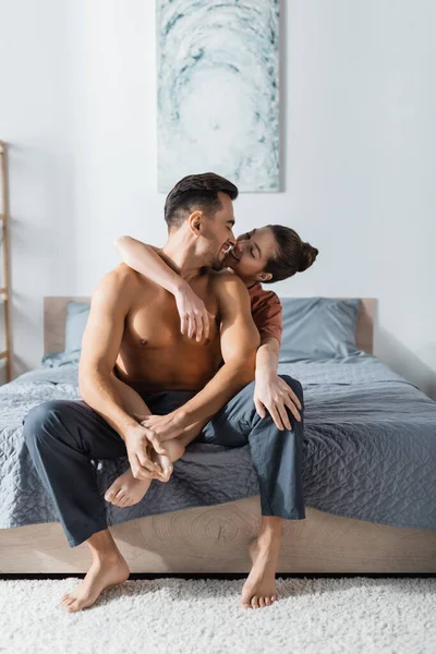 Happy and sexy woman hugging shirtless boyfriend sitting on bed in pajama pants — Stock Photo