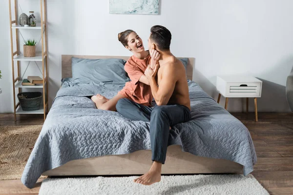 Aufgeregte Frau im T-Shirt neben hemdlosem Mann in Pyjamahose auf Bett sitzend — Stockfoto