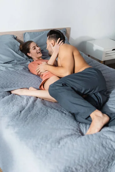 Cheerful woman hugging shirtless man in pajama pants on bed at home — Stock Photo