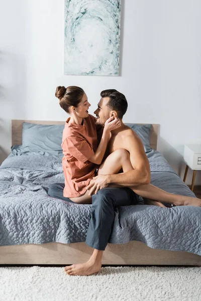 Vue latérale de la femme en t-shirt embrassant avec homme torse nu en pyjama pantalon sur le lit à la maison — Photo de stock