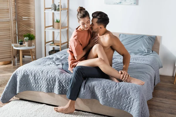 Hemdloser muskulöser Mann umarmt Freundin im T-Shirt, während er auf dem Bett sitzt — Stockfoto