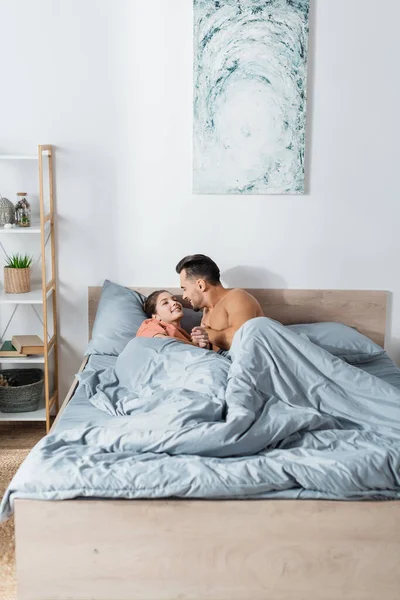 Jeune couple sexy tenant la main et se souriant tout en étant couché sous une couverture grise — Photo de stock