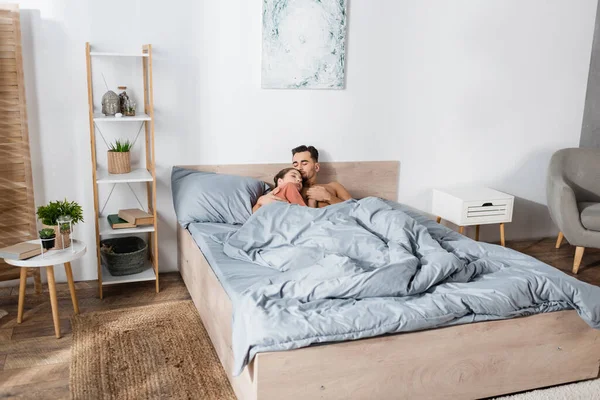 Giovane coppia innamorata che si tiene per mano mentre dorme sotto una coperta grigia nella camera da letto moderna — Foto stock