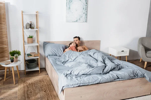 Pareja joven enamorada cogida de la mano mientras duerme bajo una manta gris en un dormitorio moderno - foto de stock