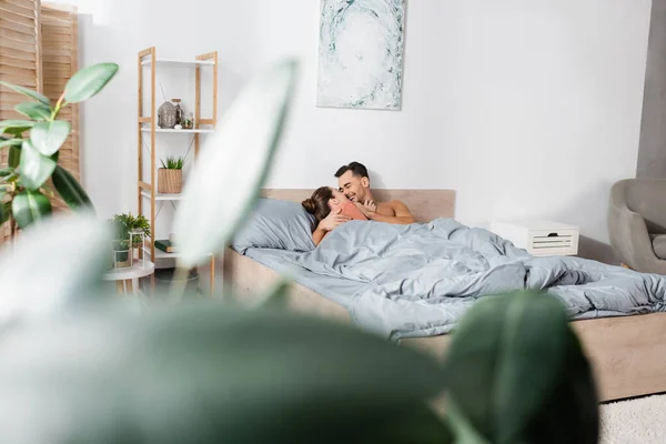 Felice giovane coppia che abbraccia e sorride sotto la coperta grigia in camera da letto moderna in primo piano sfocato — Foto stock