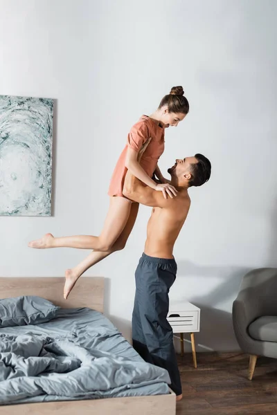 Боковой вид на мужчину без рубашки в пижамных штанах, поднимающего изумленную девушку над кроватью дома — стоковое фото