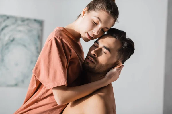 Young woman in t-shirt hugging shirtless boyfriend holding her at home — Stock Photo