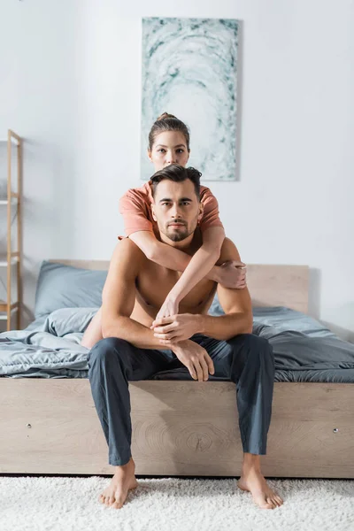 Young woman looking at camera while embracing shirtless man sitting on bed in pajama pants — Stock Photo