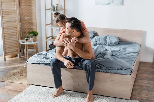 Happy shirtless man sitting on bed in pajama pants near girlfriend embracing him in bedroom — Stock Photo