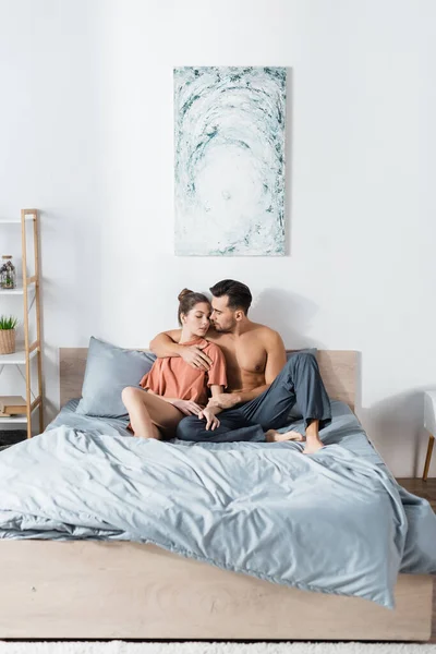 Sexy muscular man in pajama pants hugging girlfriend in t-shirt on bed at home — Stock Photo