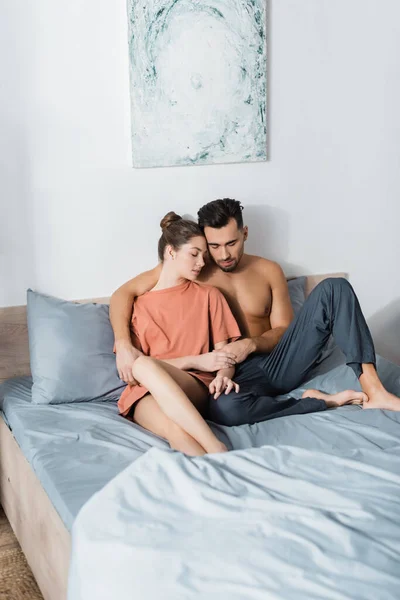 Mujer joven en camiseta y hombre sin camisa en pantalones de pijama abrazándose y abrazándose en la cama con los ojos cerrados - foto de stock