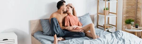 Hombre en pantalones de pijama abrazando y besando novia en dormitorio moderno, bandera - foto de stock