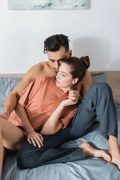Мужчина без рубашки в пижамных штанах и соблазнительная женщина в футболке, сидящая на кровати и держась за руки — стоковое фото