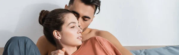 Shirtless man kissing girlfriend in t-shirt looking away in bedroom, banner — Stock Photo