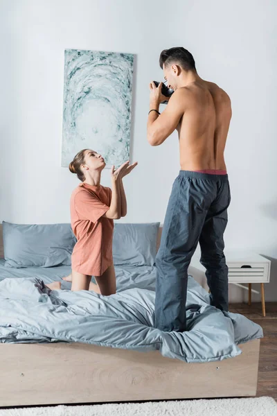 Femme sexy en t-shirt agenouillé sur le lit et souffle baiser d'air près de copain torse nu avec caméra vintage — Photo de stock
