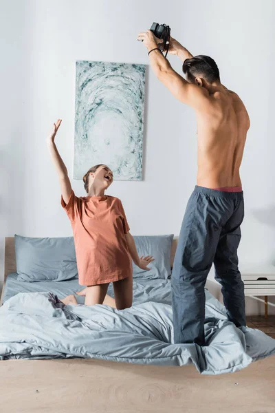 Femme excitée posant avec la main levée tout en s'agenouillant sur le lit près de petit ami avec caméra vintage — Photo de stock