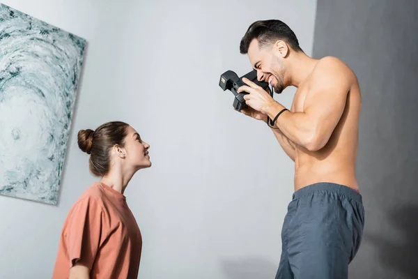 Вид збоку веселий без сорочки чоловік з старовинною камерою фотографія дівчини в футболці — стокове фото