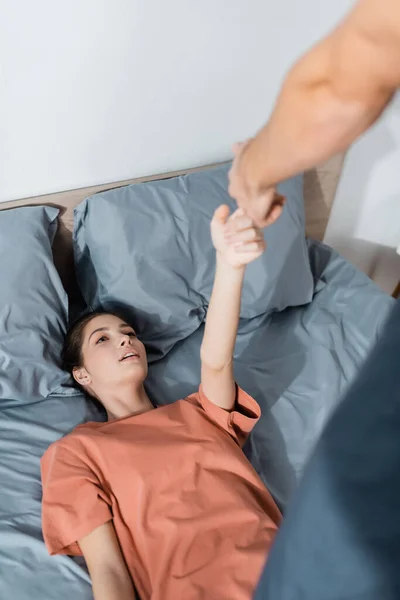 Alta vista angular de la mujer tocando la mano del hombre de pie sobre ella en la cama - foto de stock