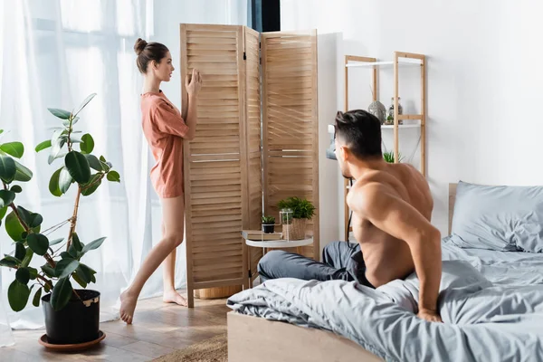 Femme sexy en t-shirt posant près diviseur de chambre et homme torse nu avec caméra vintage dans la chambre — Photo de stock