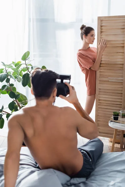 Mulher sedutora posando perto divisor de quarto enquanto homem sem camisa com câmera vintage tirar foto em primeiro plano borrado — Fotografia de Stock