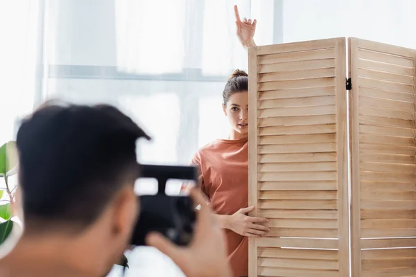 Junge Frau zeigt mit dem Finger nach oben, während sie in der Nähe von Raumteiler posiert und verschwommener Freund mit Vintage-Kamera — Stockfoto