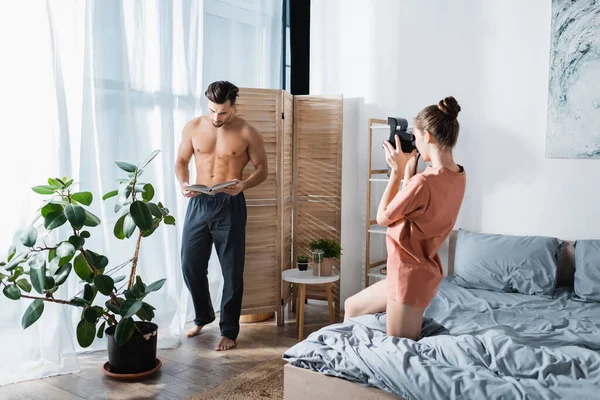 Homem muscular em calças de pijama posando com livro perto da namorada com câmera vintage — Fotografia de Stock