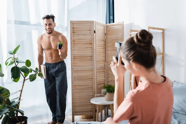 Fröhlicher Mann ohne Hemd mit Buch und Blumentopf posiert neben verschwommener Freundin mit Vintage-Kamera — Stockfoto