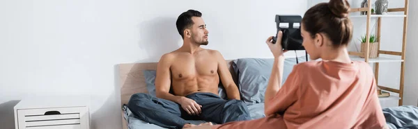 Mujer joven con cámara vintage tomando fotos de novio sexy sentado en la cama y mirando hacia otro lado, pancarta - foto de stock