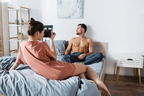 Joven mujer tomando foto de sexy novio sentado en la cama y mirando hacia otro lado - foto de stock