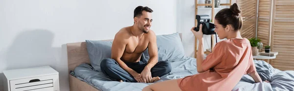 Mujer joven con cámara vintage tomando la foto del hombre sonriente sentado en la cama en pantalones de pijama, pancarta - foto de stock