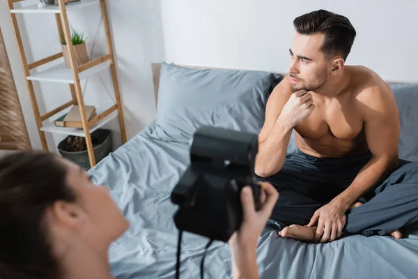 Blurred woman with vintage camera taking photo of pensive shirtless man sitting on grey bedding — Stock Photo