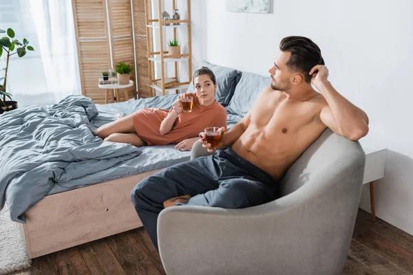 Mujer joven acostada en la cama con taza de té y mirando a su novio sin camisa en el sillón - foto de stock