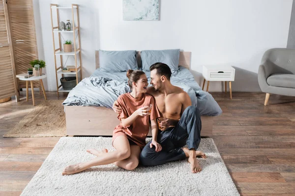 Alegre pareja con tazas de té sentados en el suelo en el dormitorio y sonrientes el uno al otro - foto de stock