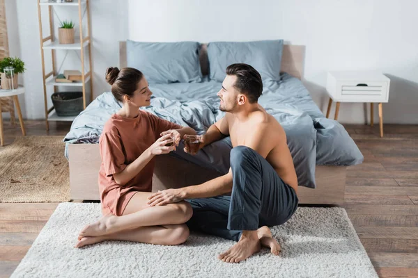 Feliz pareja joven con tazas de té hablando en alfombra de piso en el dormitorio - foto de stock