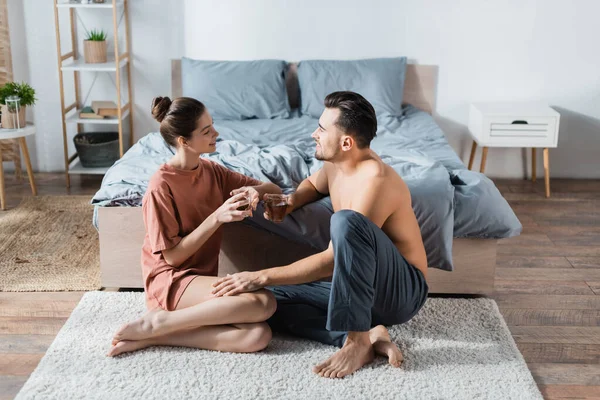 Felice giovane coppia con tazze da tè parlando sul tappeto pavimento in camera da letto — Foto stock