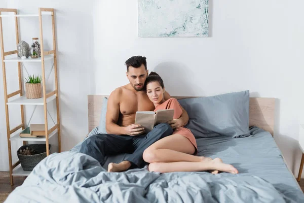 Joven y sexy pareja lectura libro en gris ropa de cama en casa - foto de stock